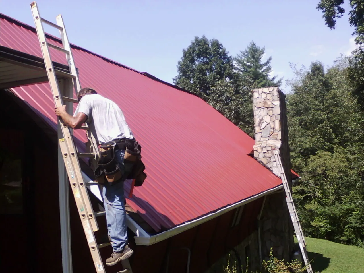 Roofer on a ladder beside a red metal roof and stone chimney — Soffit & Fascia Repair by Roofing Brothers in Delphi
